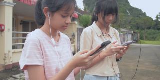 两个可爱的亚洲少女戴着耳机，站在房子外面，喜欢用智能手机聊天和看社交媒体。少女的现代生活。