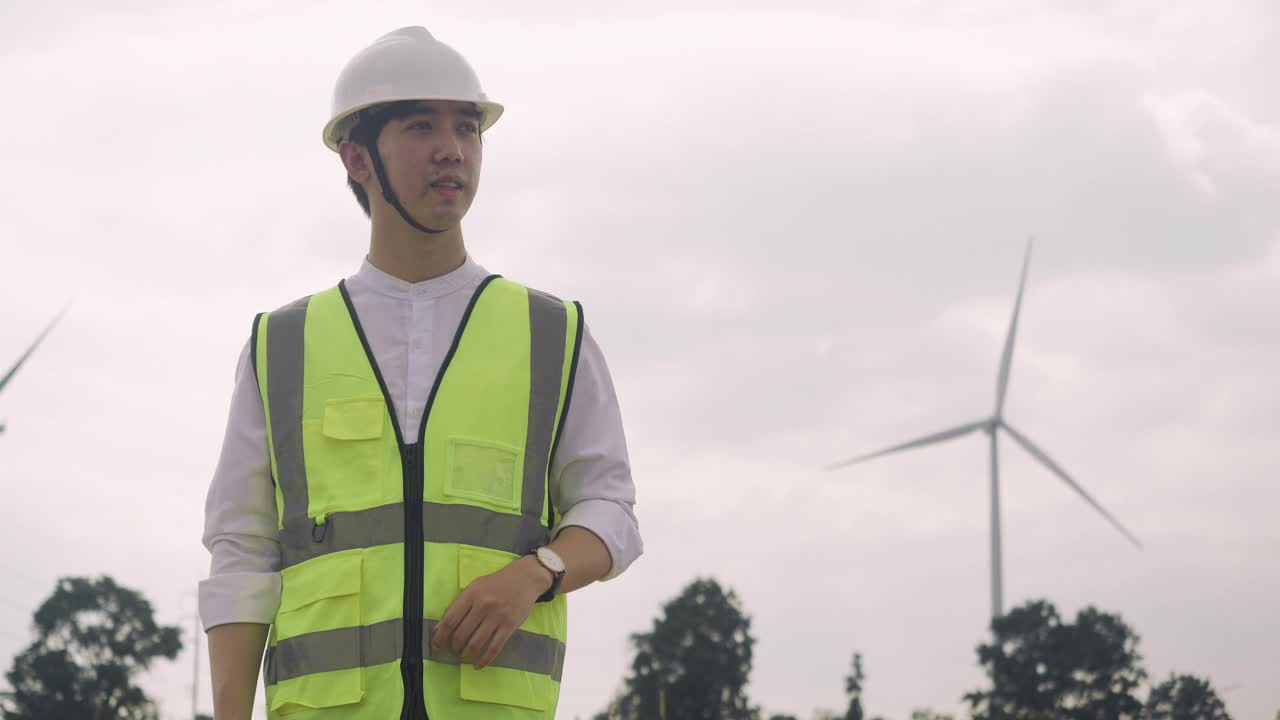 风力发电机站技术员工程师。亚洲男性工程师和女商人穿着反光夹克和头盔走在风力涡轮机农场。男老板正在用对讲机工作。