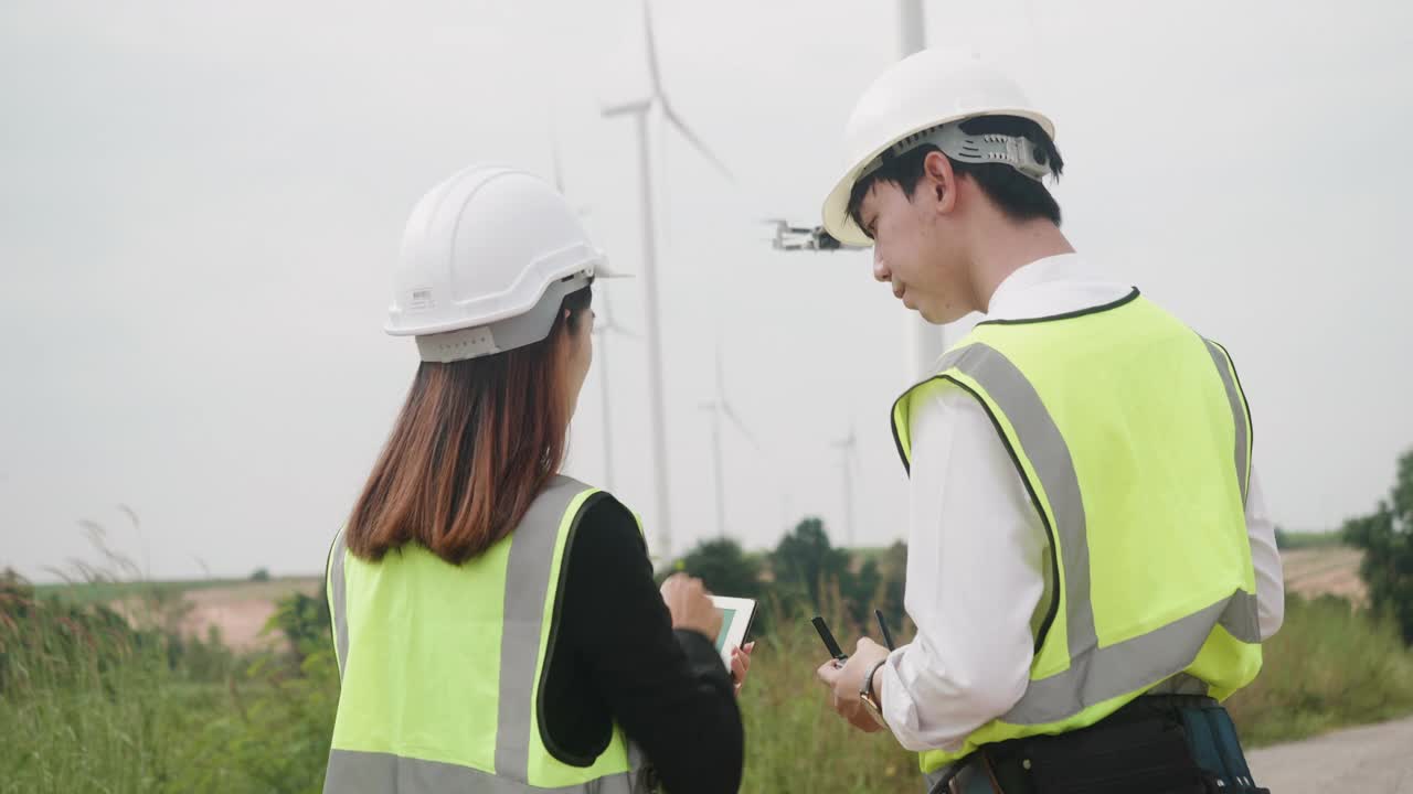 站在风力发电场的工程师，穿着反光背心和头盔的亚洲男性工程师和女商人。工程师们正在使用平板电脑和无人机工作。后视