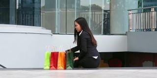 一个年轻的女人坐在购物中心前，休息着，看着她在购物袋里买的东西。