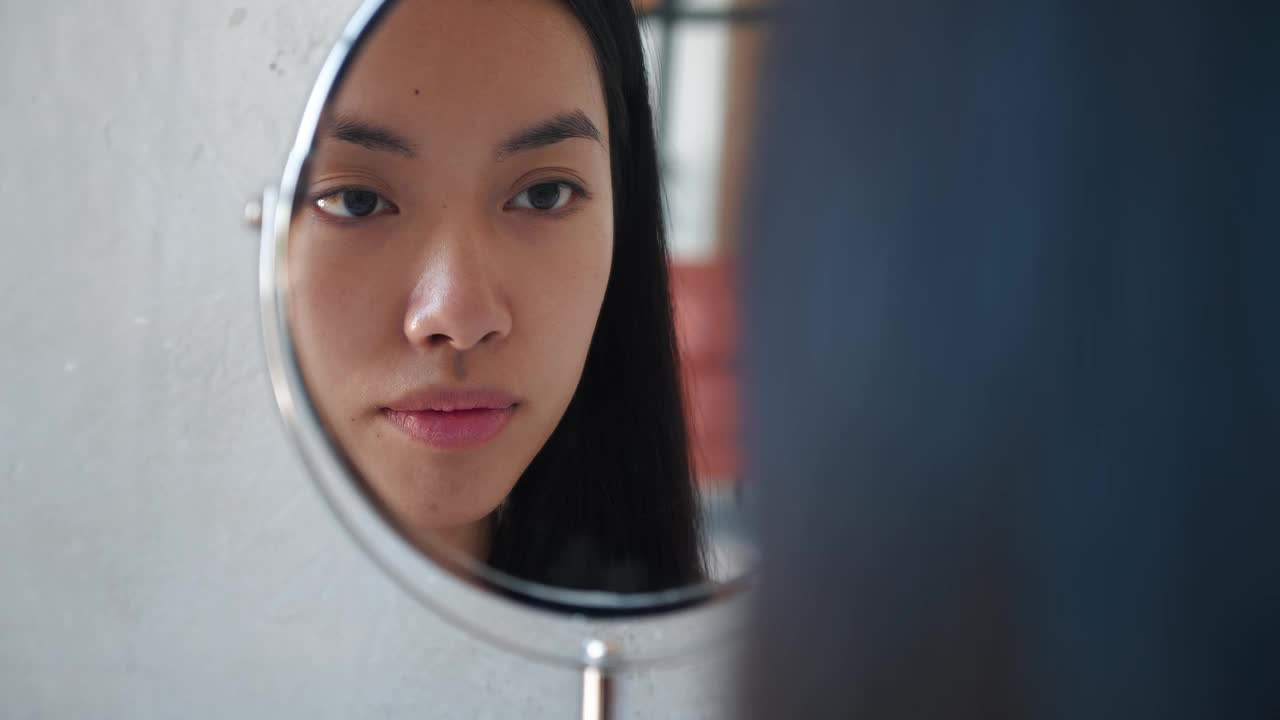 可爱美丽自然黑发的亚洲女孩用棉棉洗脸微笑着看着自己在化妆小镜中的倒影。美女用洁面乳和化妆棉卸妆