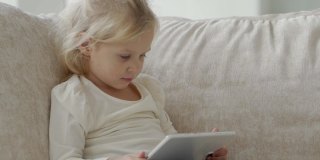 小女孩坐在沙发上看卡通片或浏览互联网时，正在使用电子平板电脑