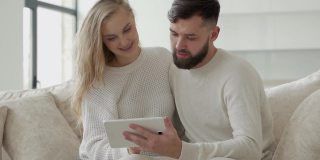 年轻幸福的夫妇，看着平板电脑屏幕，在舒适的沙发上放松。一对恋爱中的情侣在网上商店购物