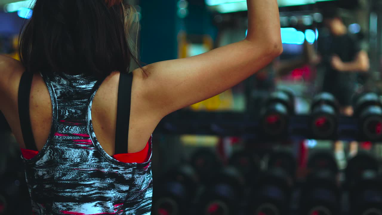 后视镜:年轻的亚洲女性在健身房里，在镜子前举起哑铃，锻炼肌肉(背部和二头肌)，锻炼力量，身体健康和结实。