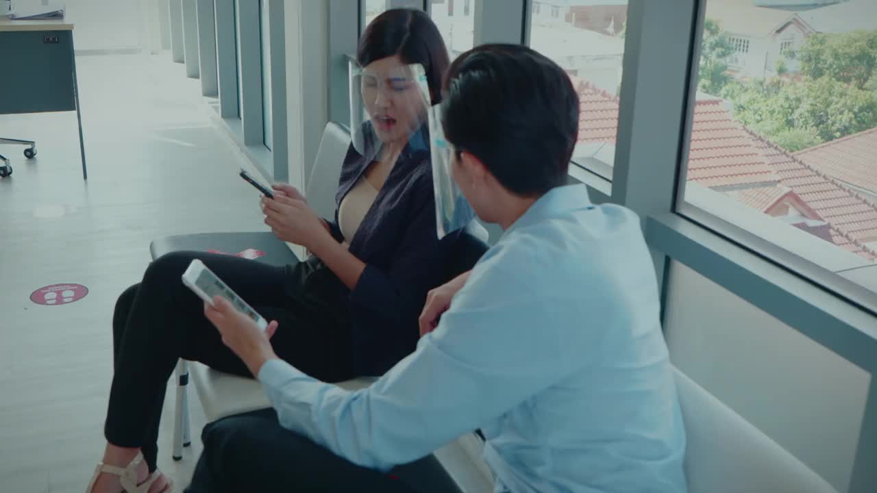 在冠状病毒大流行期间，两名亚洲同事在办公室用智能手机与客户交谈，使用面罩、互联网、技术概念、4K分辨率、社交距离。