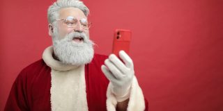 现代圣诞老人。快乐的圣诞老人正在讲电话，通过视频通话，在远处，他说你好，祝你圣诞快乐。把雪吹进电话里。红色的背景。慢动作