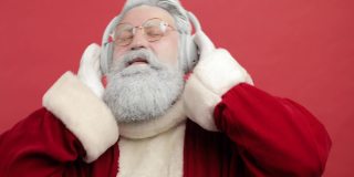 时髦开朗的圣诞老人戴着耳机听音乐，在红色的背景下唱歌跳舞。圣诞精神，圣诞晚会结束