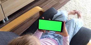 孩子坐在家里的椅子上使用智能手机，智能手机的绿色屏幕模型是水平景观模式。使用手机、浏览互联网、观看内容、视频、写博客、玩游戏