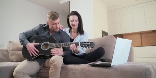 年轻夫妇坐在家里的沙发上用笔记本电脑在线学习弹吉他