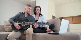 年轻夫妇坐在家里的沙发上用笔记本电脑在线学习弹吉他