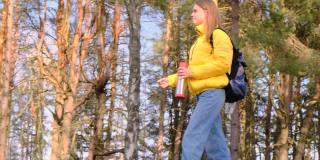 搭便车旅行的概念。一名年轻的女游客背着黄色夹克的背包，手里拿着保温瓶，沿着松树林走着。有选择性的重点。慢镜头,4 k