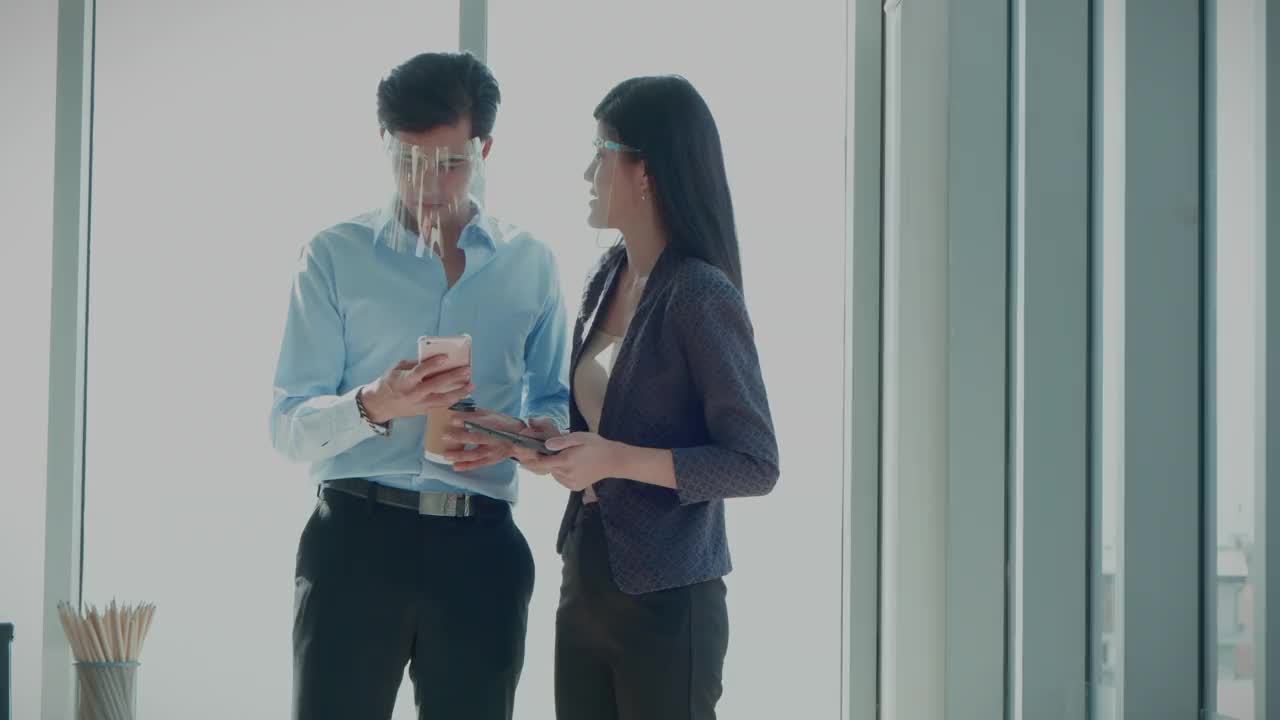 在冠状病毒大流行期间，两名亚洲同事在办公室用智能手机与客户交谈，使用面罩、互联网、技术概念、4K分辨率、社交距离。
