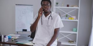 聪明的非洲裔美国人，戴着眼镜，穿着医生长袍，站在室内的餐桌旁打电话。智慧的专业医生的肖像建议治疗。医药概念。