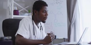 侧面观点自信的非洲裔美国科学家在网上开会，坐在桌子旁，背景是笔记本电脑和白板上的化学公式。严肃的专家化学家说话说服同事。