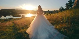 美丽的新娘跑向夕阳下的湖。Moranrange的日落和光线在背光上。穿着雪纺长裙的年轻女子