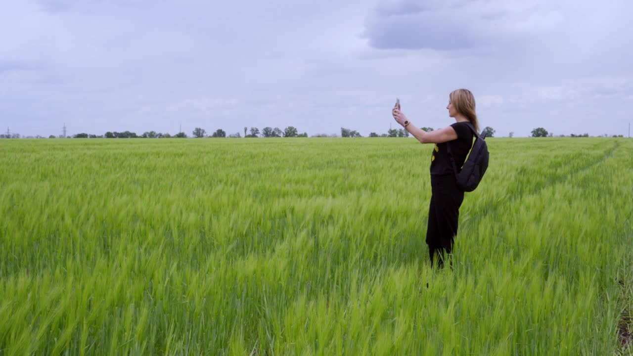 一名女游客在乡间徒步行走，站在一片生长着小麦的绿色田野上，用手机拍照。