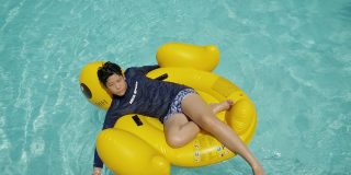 快乐的亚洲男孩放松在黄鸭橡皮筏在游泳池与家人度假，生活理念。