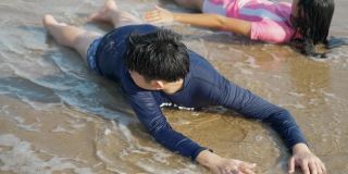 快乐的亚洲孩子们穿着泳衣一起躺在阳光明媚的沙滩上，慢动作。