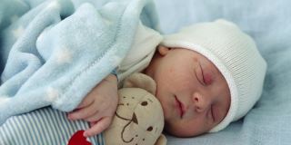 新生儿脸部特写，早期与泰迪熊睡觉的蓝色背景。在帽子上的生命开始分钟的孩子。婴儿，分娩，出生的最初时刻，开始的概念