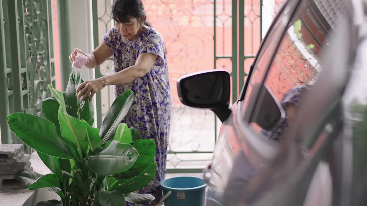 亚洲退休的老年妇女在她的家前面的一个小花园用回收塑料杯作为浇水罐浇灌植物叶子树，积极的老年生活方式的概念，侧视图