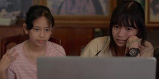 两个亚洲少女在家里用笔记本电脑从远程老师那里学习在线课程。泰国少女们坐在一起，戴着耳机在电脑屏幕上看电影。