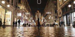 一个穿着米色长大衣、戴着围巾的千禧女孩走在冬日米兰的夜街上。穿暖和衣服的女人