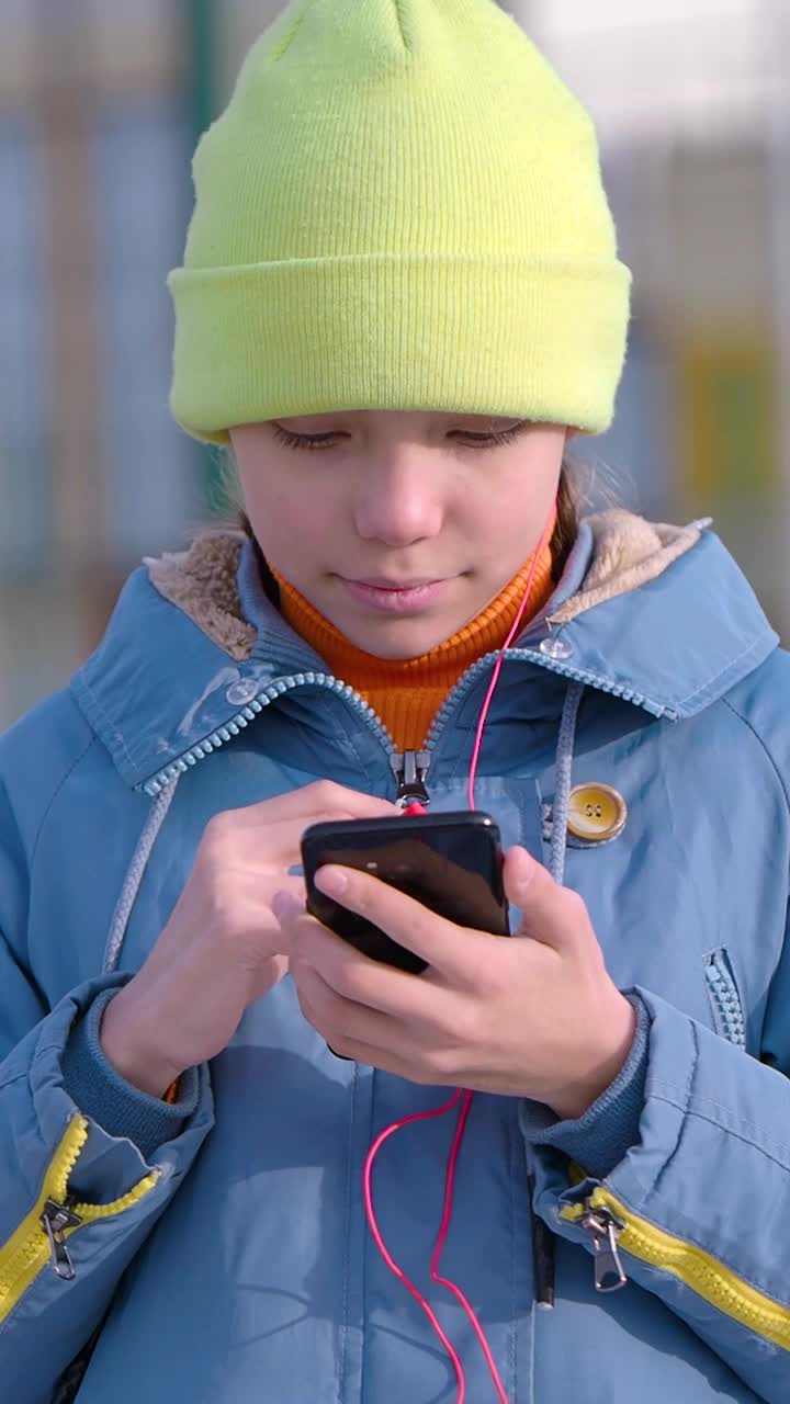 垂直视频——一个十几岁的女孩在智能手机应用程序中选择音乐，把耳机放在耳朵里欣赏音乐