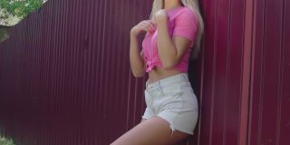 金发年轻女子粉红色背心和牛仔短裤放松在深红色铁栅栏。美丽的女孩抚摸着她的头发。暑假。人们生活方式的概念。
