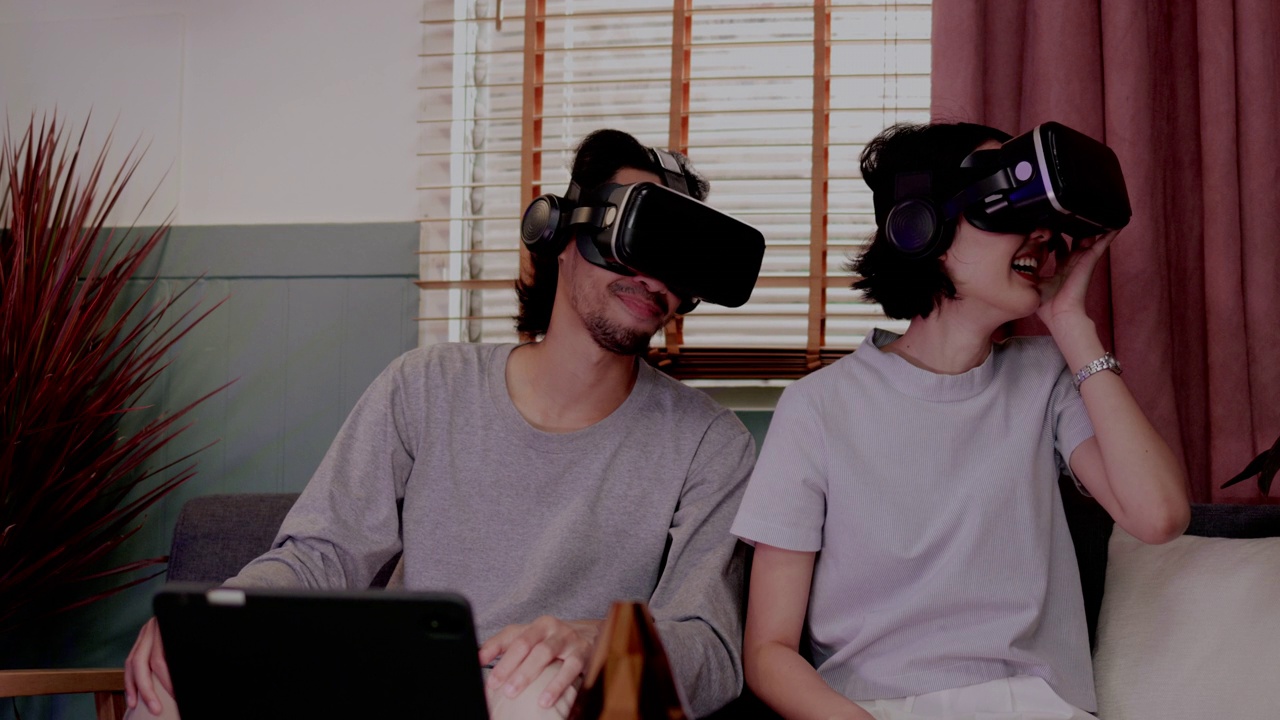亚洲夫妇戴着现代虚拟现实眼镜坐在家里的客厅里看电影。节日活动家庭娱乐技术。幸福生活在一起。metaverse概念