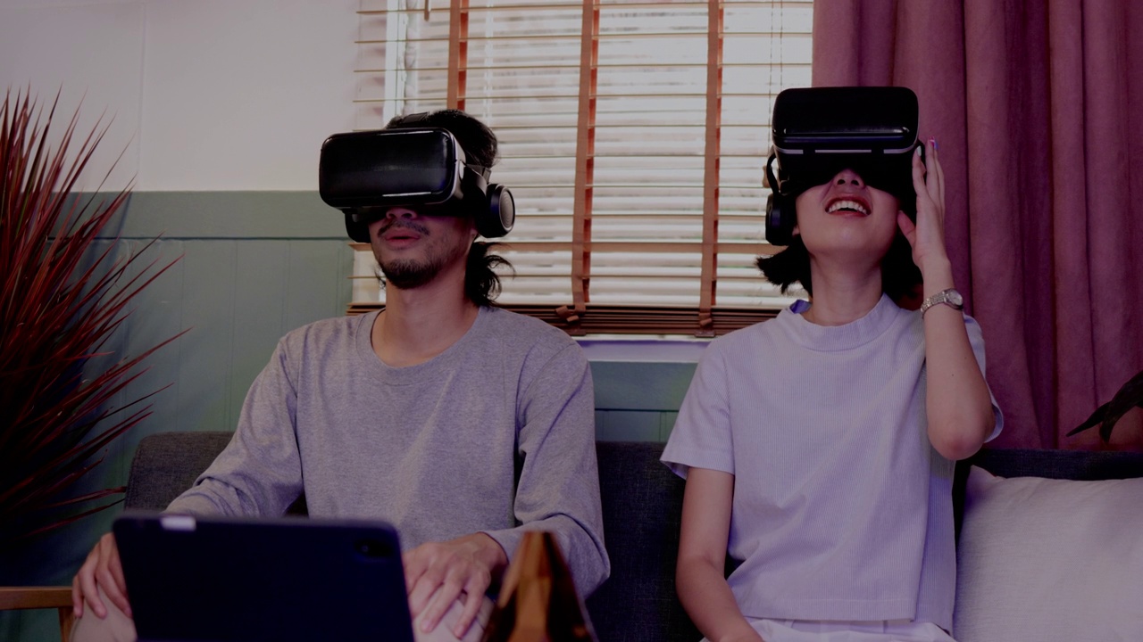 亚洲夫妇戴着现代虚拟现实眼镜坐在家里的客厅里看电影。节日活动家庭娱乐技术。幸福生活在一起。metaverse概念