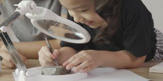 一位亚洲母亲和她的女儿正在家里通过放大镜观察化石，学习科学实验。展示了寓教于乐、充满好奇的家庭学习理念。