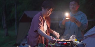 一个亚洲中国女人在晚上露营时准备烧烤，而她的丈夫用他的智能手机点亮了场景