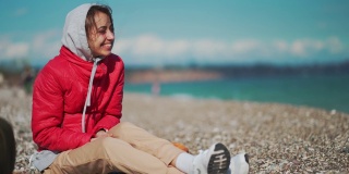 一个穿着红色夹克和连帽衫的年轻女子，在阳光明媚的多风的日子里，坐在卵石海滩上捡卵石