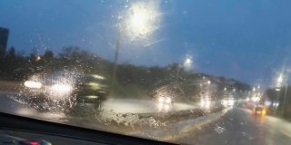 特写大雨落在汽车的挡风玻璃上，雨刷工作迅速。雨点从汽车前窗倾泻而下。开着前灯的汽车在高速公路上开会。4 k的片段