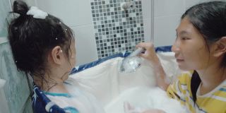 亚洲女孩淋浴她的姐姐的脸，同时放松在塑料浴缸一起在家里，生活方式的概念。