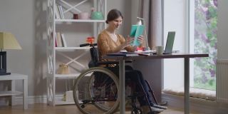 宽镜头的照片，微笑的美丽的年轻女子在轮椅挥手，在平板电脑视频聊天，坐在桌子在室内。快乐自信迷人的白人女士在网上聊天慢动作。