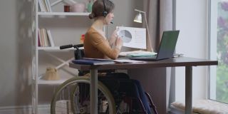 自信的白人年轻女子的肖像在轮椅上，在笔记本电脑视频聊天指着图表。美丽苗条的自由职业者工作消息在线从家庭办公室。远程工作的自由。