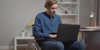 一名自信而专注的年轻残疾男子在笔记本电脑键盘上发短信，看着相机微笑。满意快乐的白人自由职业者在轮椅摆姿势在家里的慢动作。