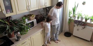 父亲和儿子抬起他们的腿，让机器人真空吸尘器清理厨房的地板