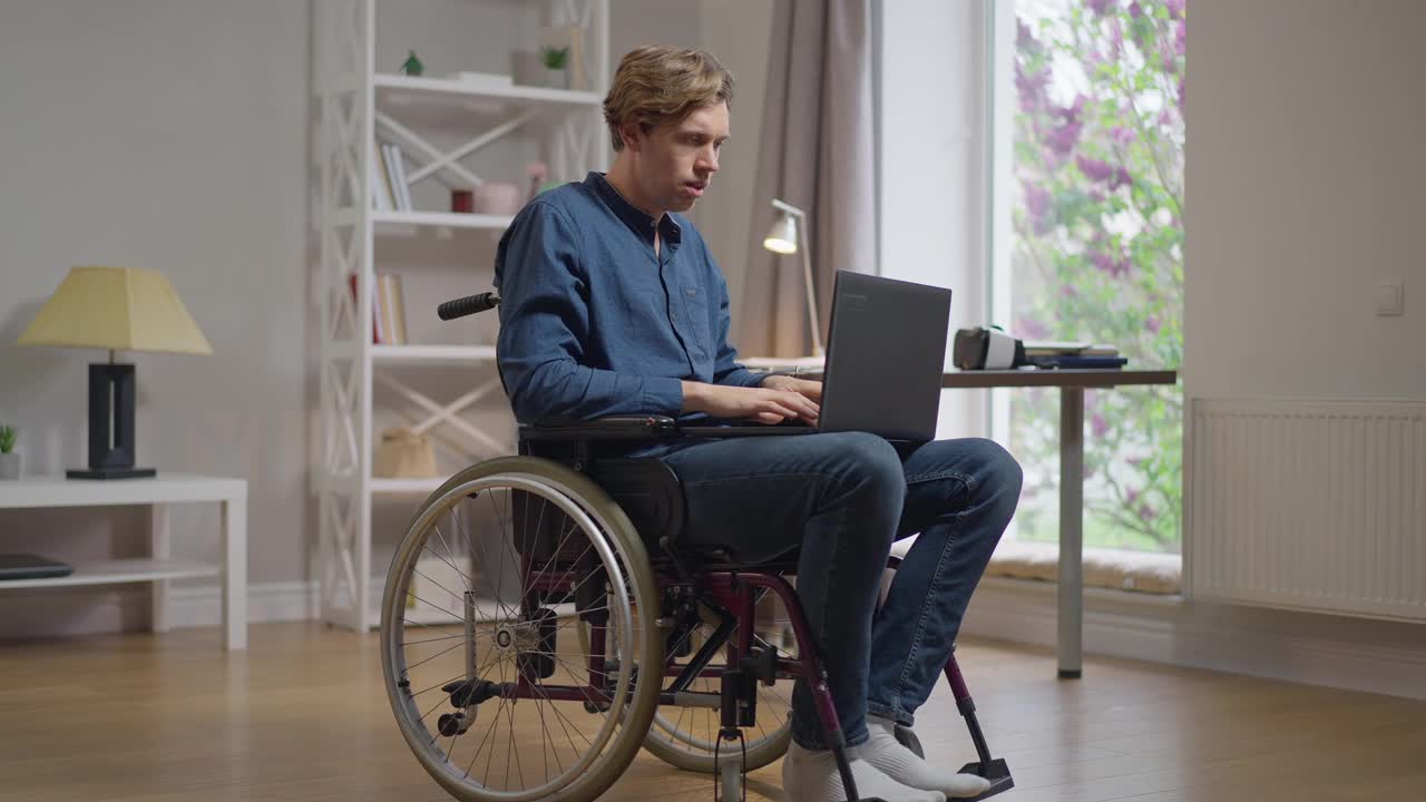 一个坐在轮椅上专注的聪明人，在网上发短信，在笔记本电脑键盘上打字，思考。集中自信的白人残疾人在家里客厅上网。