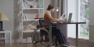 注意力集中的聪明残疾年轻妇女在轮椅上冲浪在笔记本电脑分析文件在室内的家庭办公室。宽镜头人像聪明的白人自由职业者工作在慢动作。