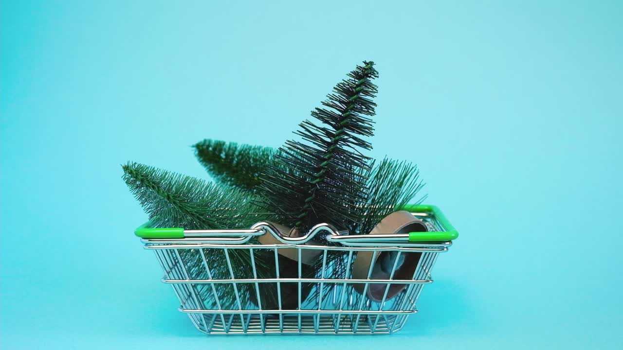 把小圣诞树放进购物车的手。蓝色背景下超市手推车上的人造迷你圣诞树。圣诞节，新年，假日购物概念。极简主义。