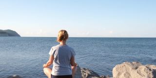 清晨，女孩坐在海边的石头上练习瑜伽。日出时，一位年轻女子坐在海边的莲花位上冥想。自然、健康、个人活动的概念