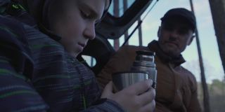 爸爸和十几岁的儿子一起坐在敞开的汽车后备箱，喝着热茶