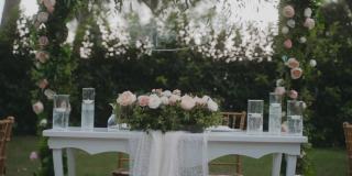 令人惊叹的婚礼布置在花园，白色桌布和木制椅子的桌子。美丽的节日气氛。花园里到处都是粉红色和白色的玫瑰。