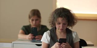 两个女学生坐在学校的课桌前，看着自己的智能手机。儿童和电子产品，青少年和手机在学校