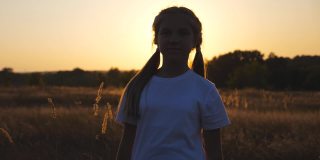 小女孩站在草地上，以夕阳为背景，对着相机微笑。快乐的小女孩玩她的头发休息在草地。自由自在的概念。多莉拍摄