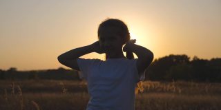 快乐的小女孩站在草地上玩着自己的头发，对着镜头。小微笑的女孩的肖像在草地上日落的背景。自由自在的概念。多莉拍摄