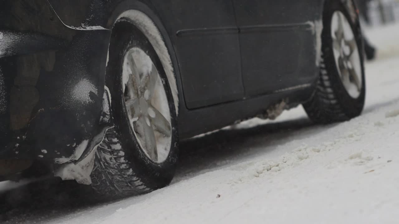 汽车正站在冬天城市里白雪覆盖的十字路口。排气管中冒出的烟雾。降雪。车轮的慢动作特写。轮胎。交通状况不佳，暴风雪。危险之旅