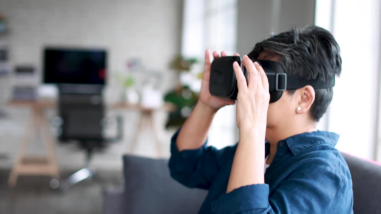 一位年长的亚洲老妇坐在家里的客厅里玩完虚拟现实眼镜，一放就感到兴奋和满足。退休者学习新技术体验的概念。